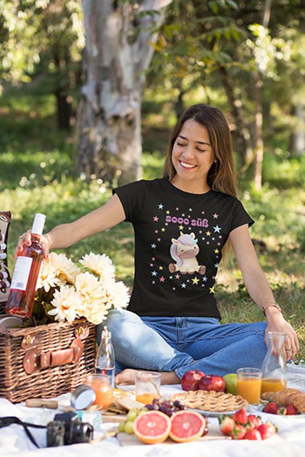 Die junge Dame sitzt auf einer Decke mit einem Picknickkorb. Sie trägt ein schwarzes T-Shirt mit einem bunten Einhorn und vielen Sternen. Auf dem T-Shirt steht sooo süß.